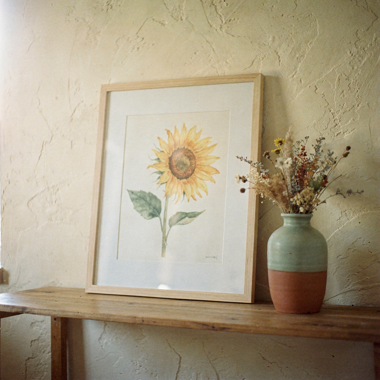 Framed sunflower botanical print leaning against a warm cream plaster wall with a ceramic vase